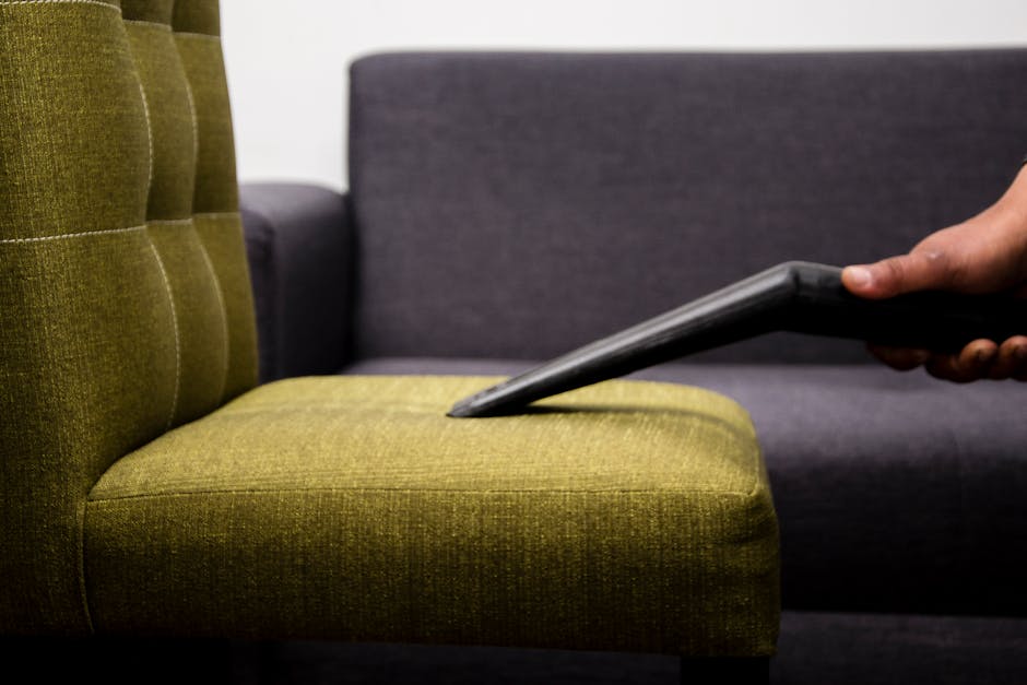 A person in a dark jacket is vacuuming a yellow and white patterned cushion placed on a dark fabric sofa in a room with neutral-colored walls, demonstrating surface cleaning and upholstery sanitisatio
