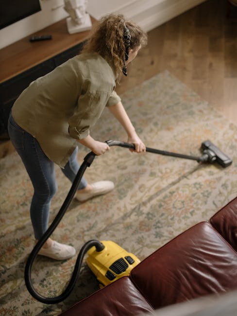 A close-up view of a person using a handheld vacuum cleaner to clean a stained olive green upholstered armchair with a tufted backrest, situated in a room with a dark grey sofa in the background and b