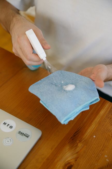 Close-up of a person's hand holding a white spray bottle, applying a cleaning solution onto a blue microfiber cloth placed on a wooden surface in a domestic setting. The background includes a portion 